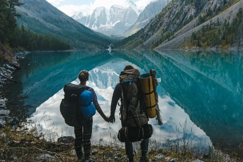 Couple with backpacks holding hands at the edge of a mirror-still mountain lake