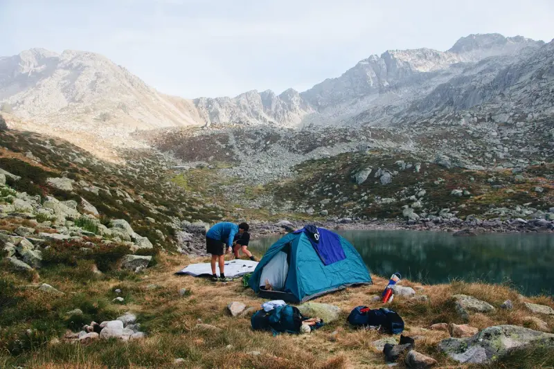 Campers pitching a blue tent beside a mountain lake in a rocky alpine valley