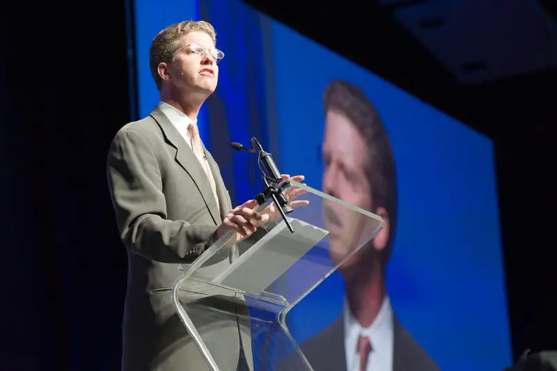 HUD Secretary Shaun Donovan speaking at a clear podium in front of a large video screen at Greenbuild
