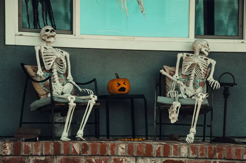 Two skeleton props seated on porch chairs beside a carved pumpkin