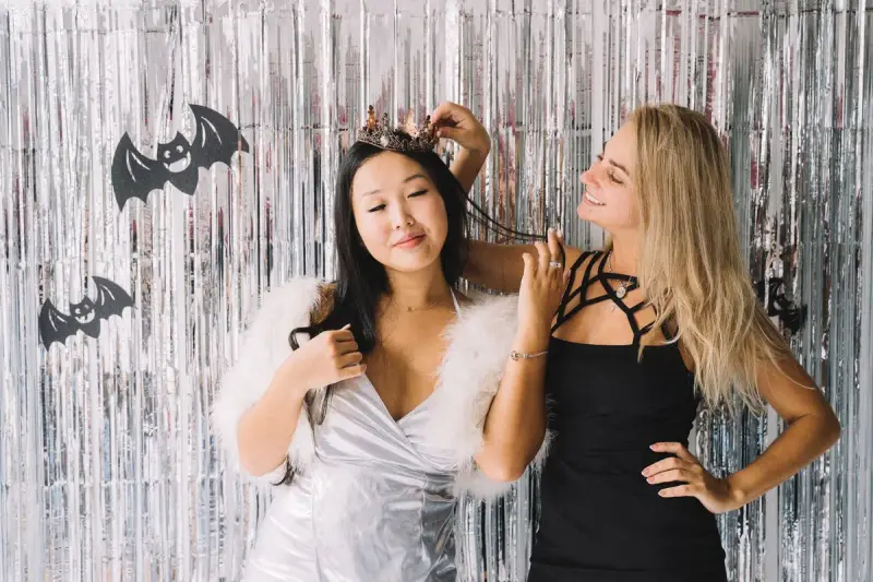 Two women in Halloween costumes adjusting a tiara in front of a silver foil curtain with paper bats