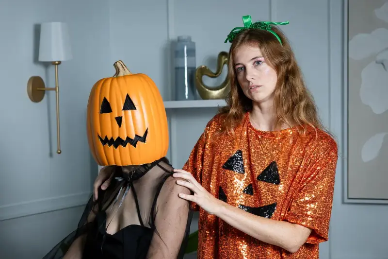Adults in Halloween costumes indoors, one wearing a carved pumpkin head