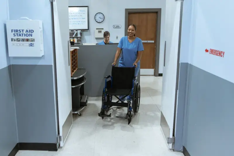 Nurse in scrubs pushing an empty wheelchair down a hospital hallway
