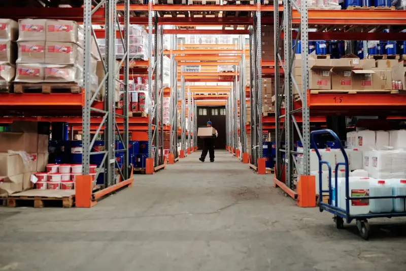 Worker carrying a carton down a warehouse aisle between stocked racks