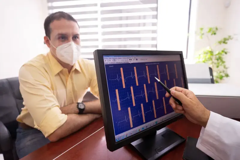 Doctor points with a pen at ECG waveforms on a screen while a seated man listens