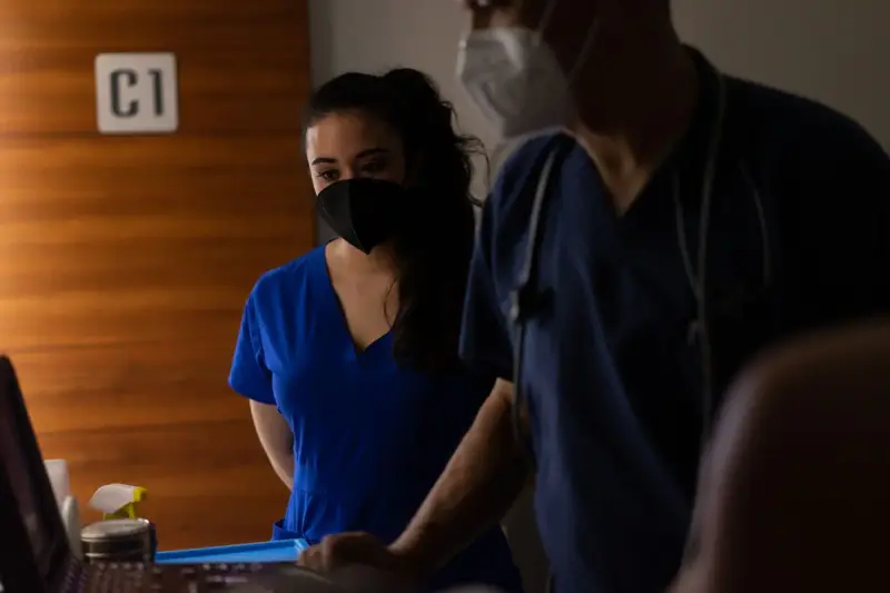 Two clinicians in blue scrubs and masks reviewing data at a workstation in a clinic room