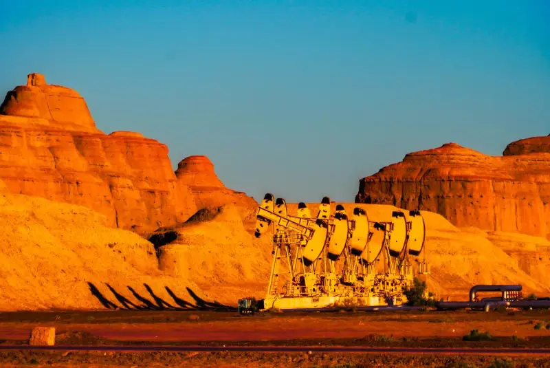 Line of pumpjacks below red rock cliffs lit by sunset