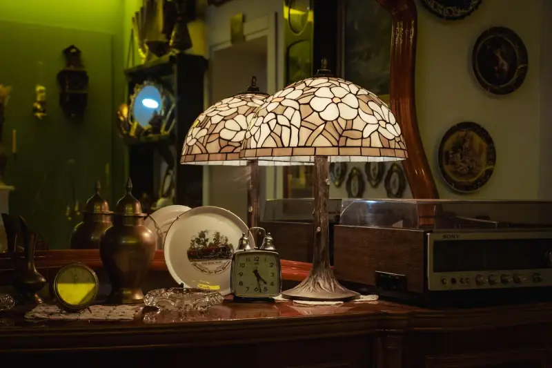 Two stained-glass table lamps glowing among plates and brass pieces on a dark wooden sideboard