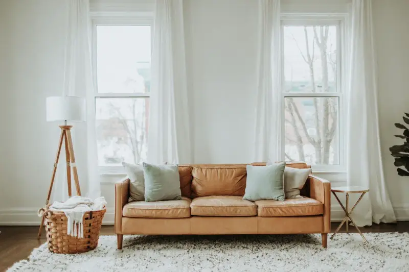 Tan leather sofa with pale cushions, a tripod floor lamp and basket in a bright living room