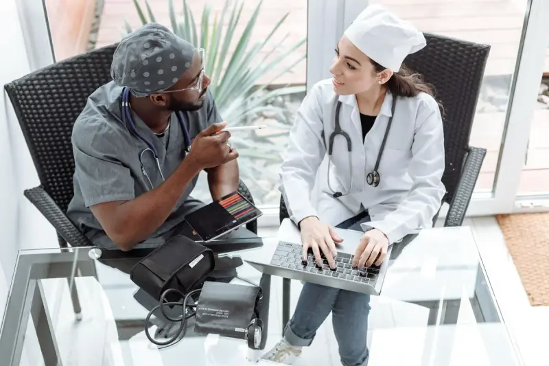 Two doctors at a glass table discussing patient care over a laptop and tablet