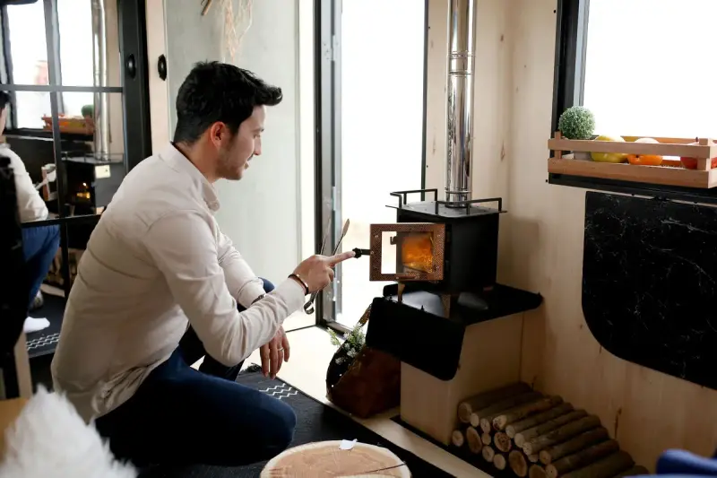 Man lighting a compact wood stove in a modern room with stacked firewood