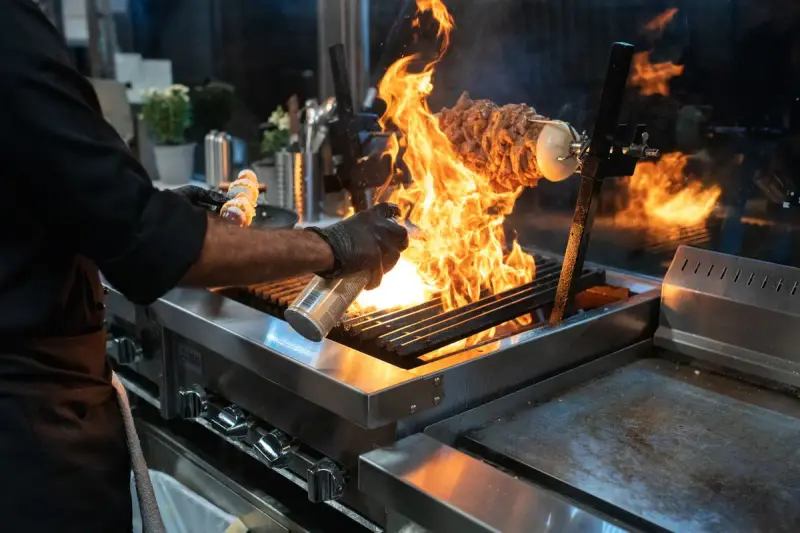 Chef grilling meat over high flames on a stainless steel grill