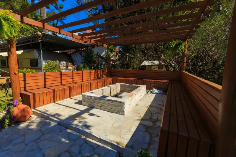 Stone fire pit with built-in wooden benches under a timber pergola on a sunny patio