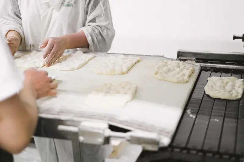 Bakers in white coats shaping flatbread dough on a conveyor belt beside a metal production line
