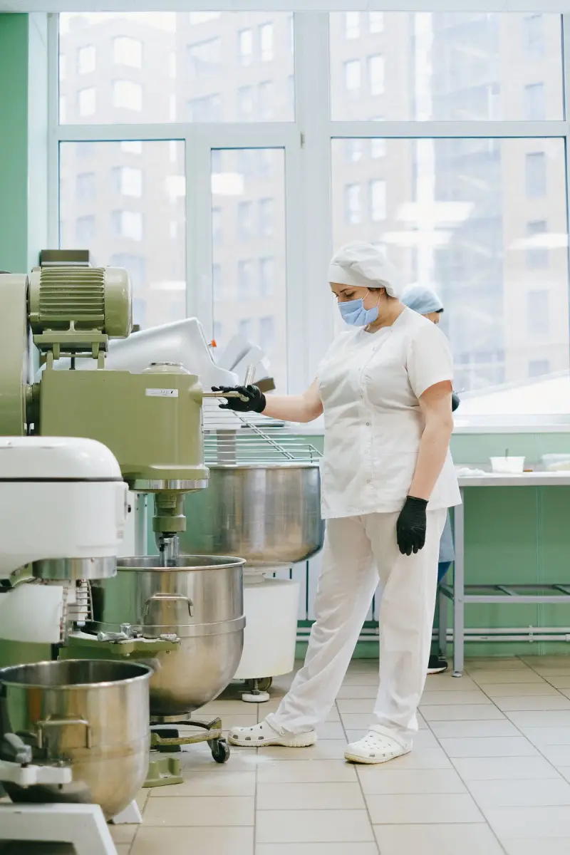 Woman in white protective clothing, mask and gloves operating large industrial dough mixers