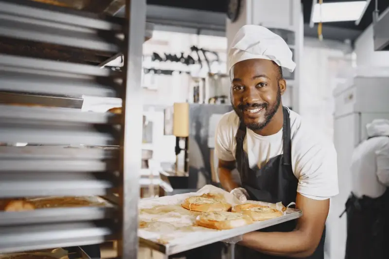 Smiling baker in a white cap holding a tray of fresh pastries beside a bakery rack