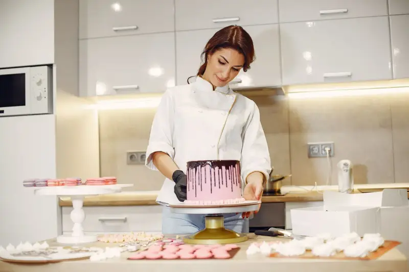 Pastry chef in a white jacket decorating a pink drip cake surrounded by macarons and meringues