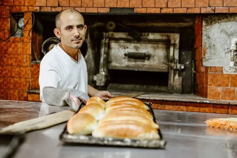 Baker in a white shirt and oven glove presenting a tray of freshly baked bread loaves