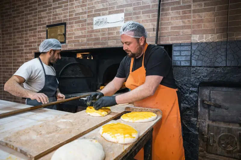 Two bakers in hairnets and aprons preparing flatbreads beside a brick oven in a traditional bakery