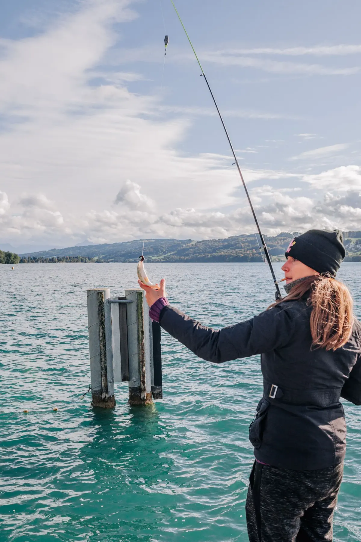 A woman fishes beside a lake with mountains in the distance.