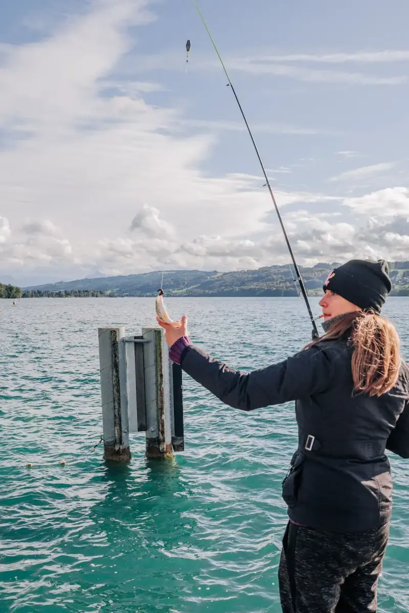 A woman fishes beside a lake with mountains in the distance.