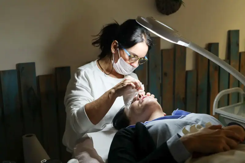 Esthetician in a mask and glasses performing a facial under a magnifying lamp
