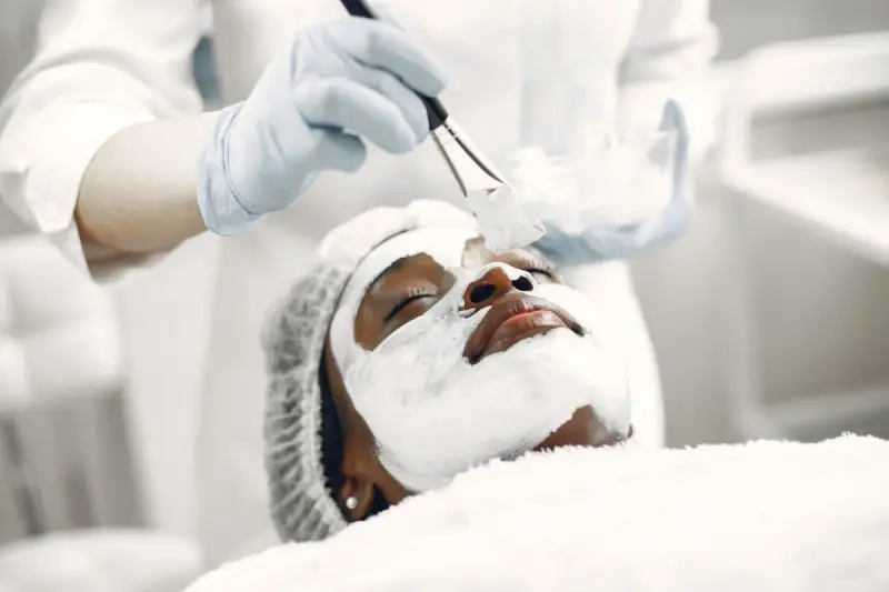 Gloved esthetician brushing a white mask onto a client's face
