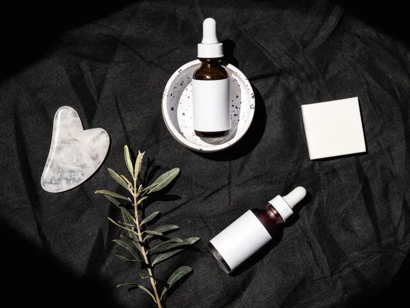 Serum bottles, a gua sha stone and a green sprig on dark linen