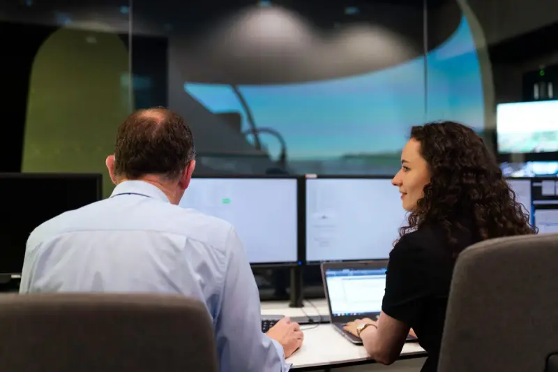 Two engineers discussing work at computer screens in a curved control room