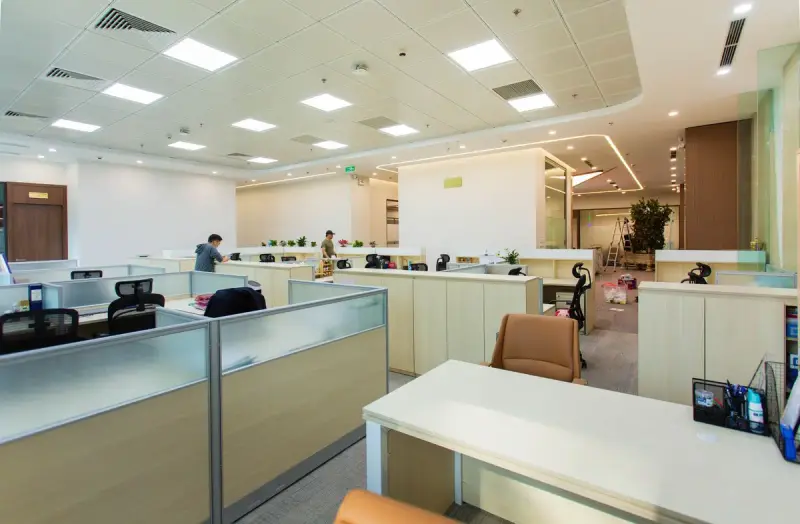Wide open-plan office with low cubicles, ceiling grid lighting and a corridor of workstations