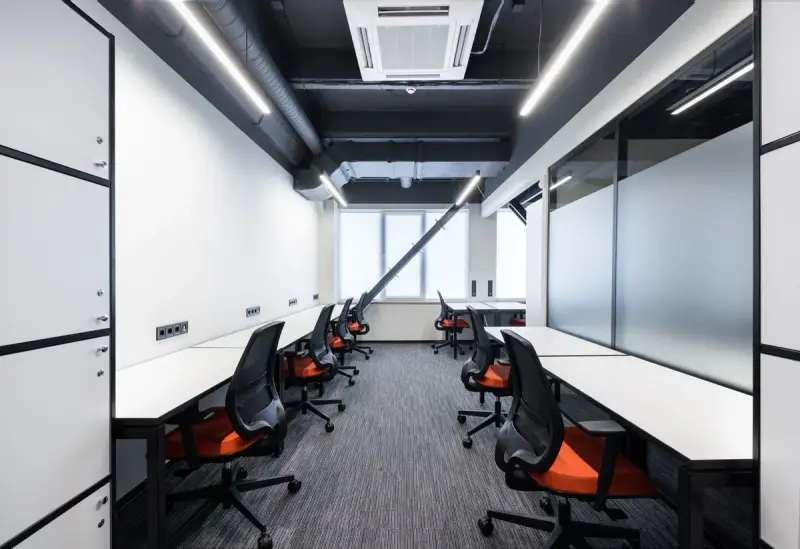 Narrow office with white desks, orange task chairs, linear lighting and a ceiling cassette unit