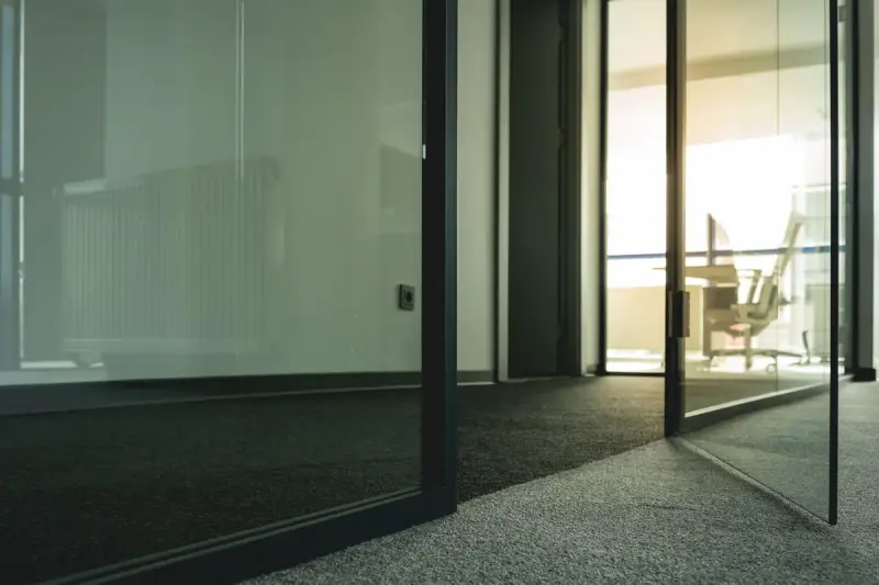 Black-framed glass office door and partition opening onto a sunlit room with carpet tile