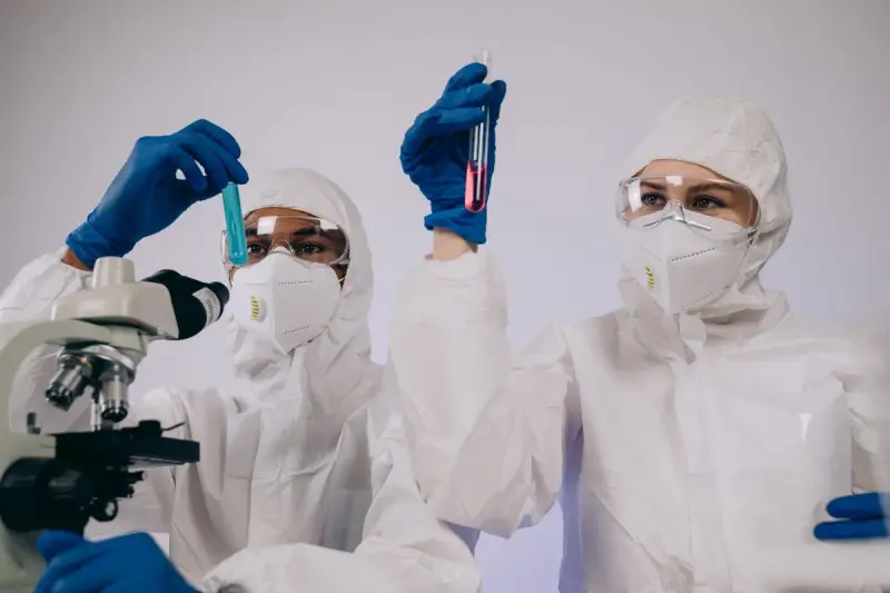 Two researchers in protective suits and goggles examining test tubes beside a microscope