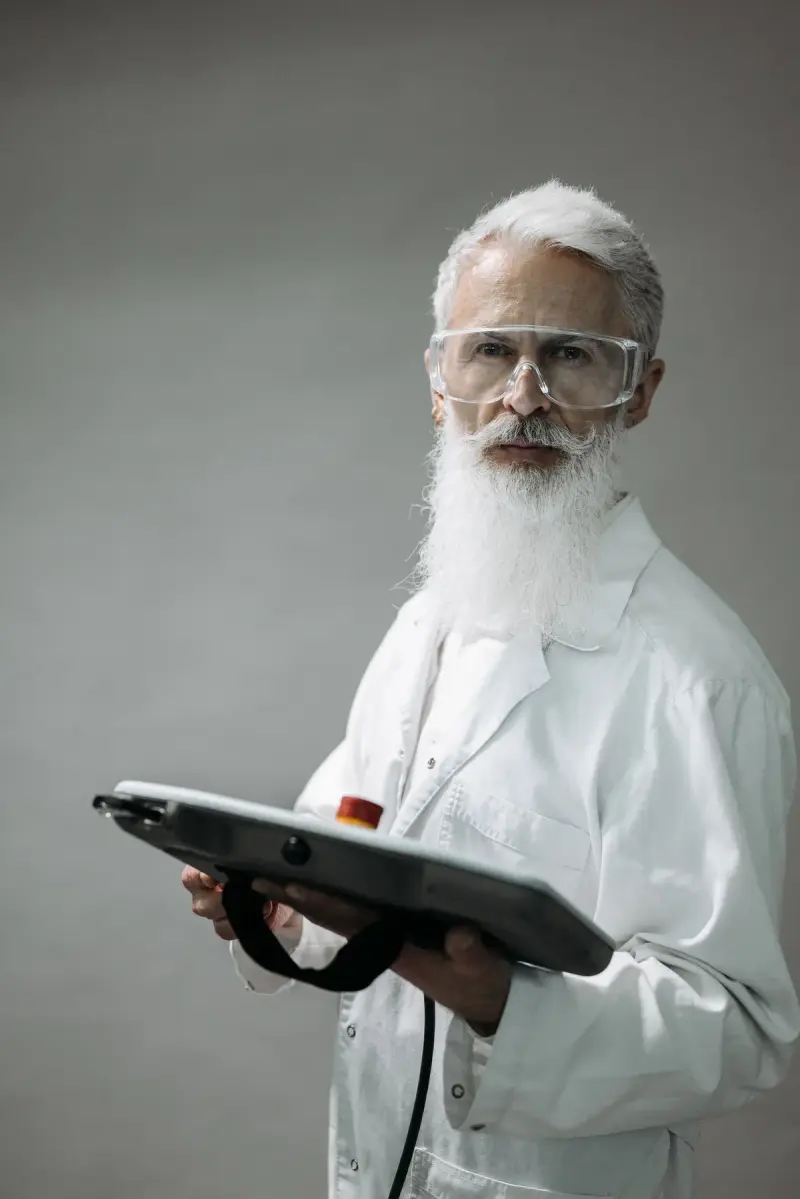 Senior scientist in a lab coat and safety glasses holding a control tablet