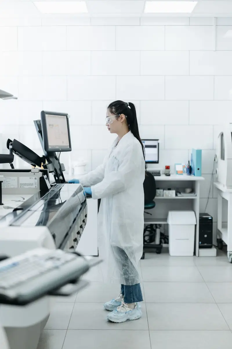Lab scientist standing at an instrument workstation with monitors in a white lab