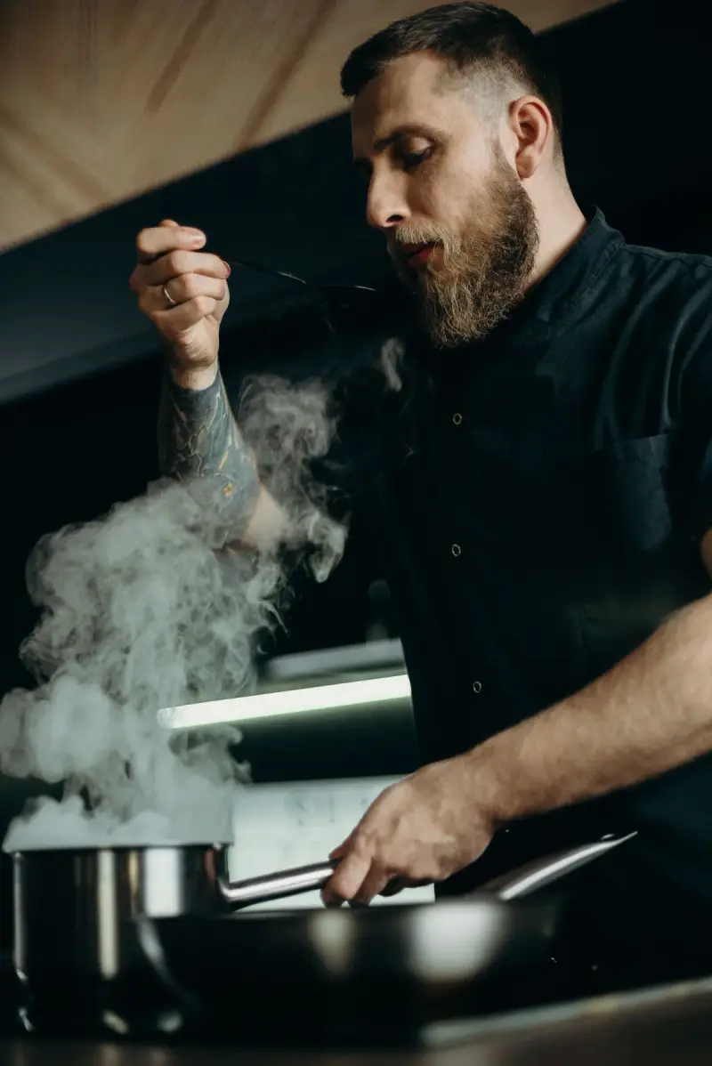 A chef tastes a dish in a professional kitchen with steam rising.