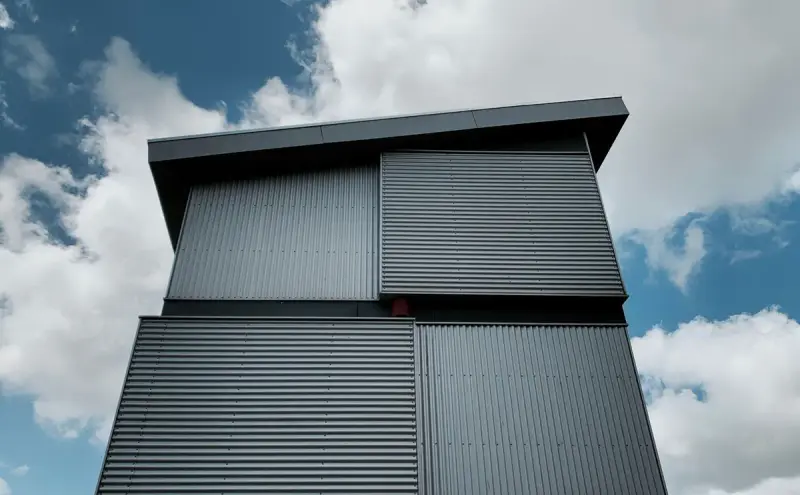Dark corrugated metal-clad building with deep overhangs against a cloudy sky