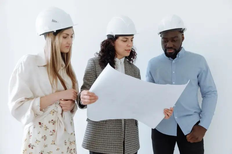 Three design professionals in white hard hats reviewing a large plan sheet
