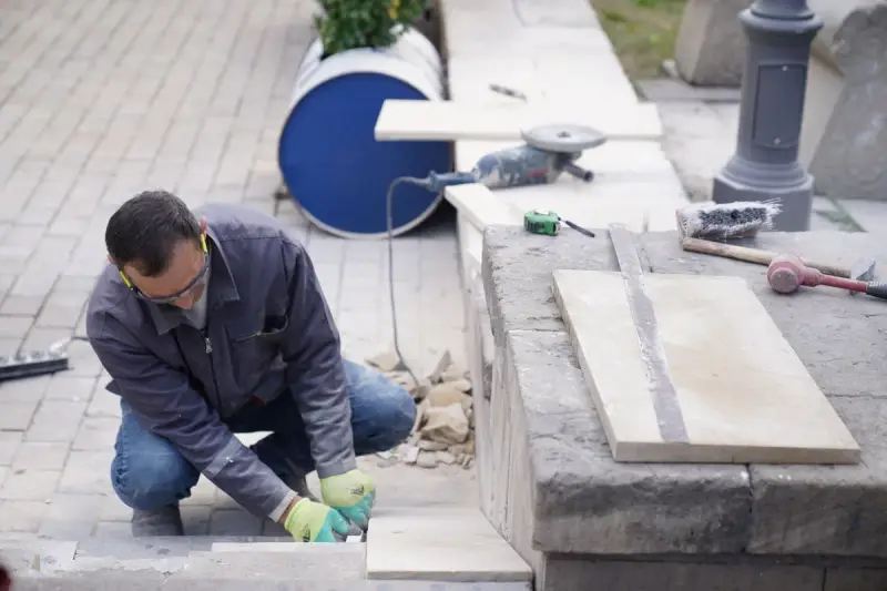 Mason in safety glasses and gloves setting stone pavers beside a seat wall