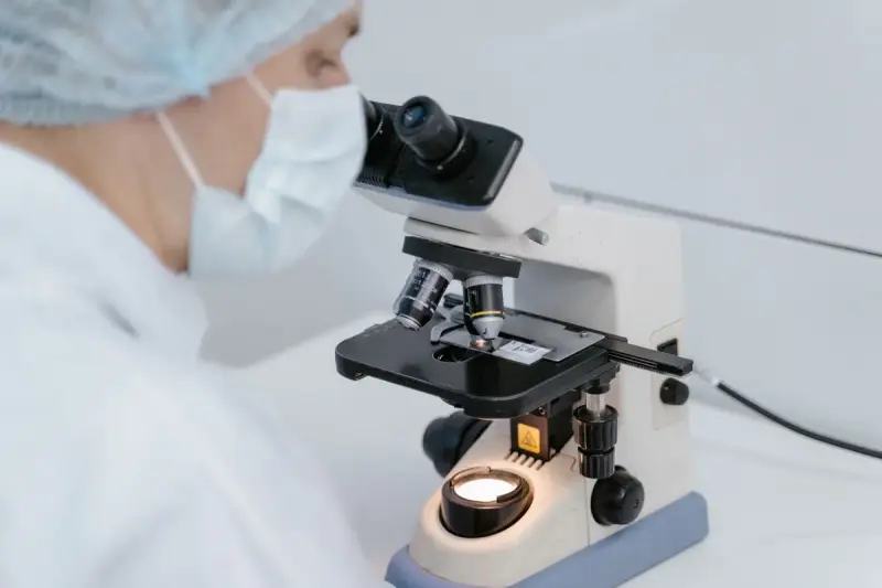 Lab worker in a hairnet and mask at a lit microscope stage