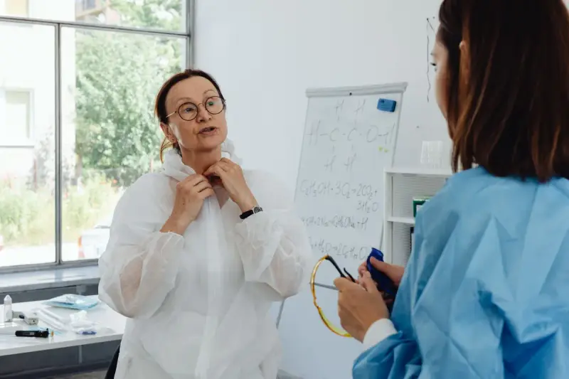 Two women researchers in lab gowns talking by a window, one in a blue protective gown