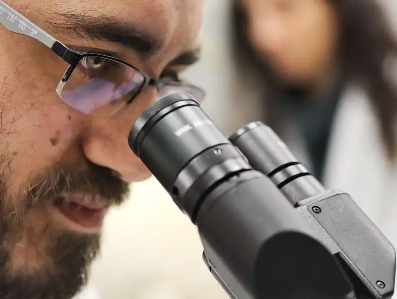 Close view of a bearded scientist in glasses looking into a microscope eyepiece