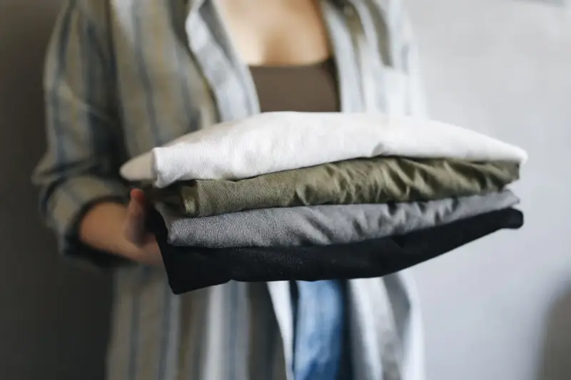 Hands holding a stack of folded t-shirts in white, olive, grey and navy
