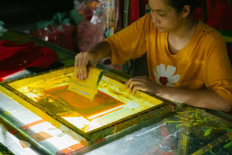 Young woman screen printing a design with a glowing yellow screen in a workshop
