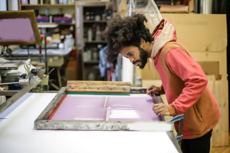 Smiling craftsman in a hoodie working a screen printing frame in his workshop
