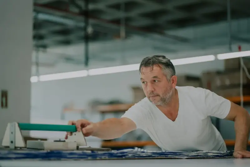 Craftsman leaning over a print table to work a squeegee across fabric