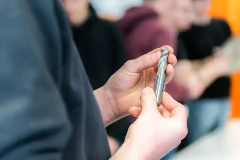 Hands holding a solid carbide end mill while a group looks on in a workshop