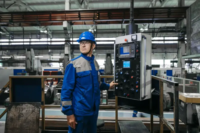 Machinist in a blue uniform and hard hat standing at a CNC control pendant on a shop floor