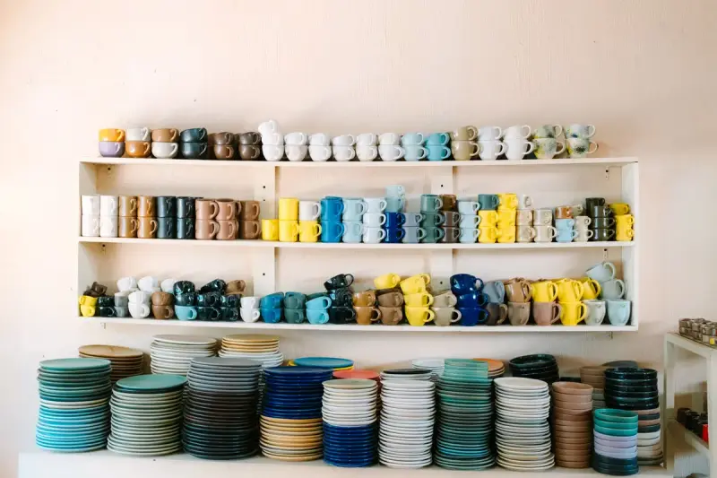 Rows of colorful ceramic mugs above stacks of glazed plates on white wall shelves