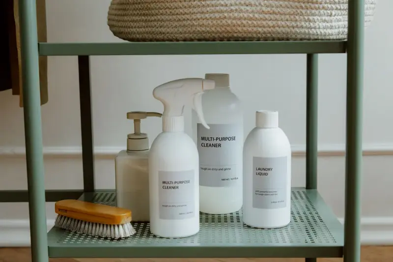 Plain white bottles of multi-purpose cleaner and a scrub brush on a green metal shelf
